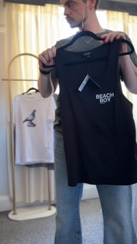 Melnā bezpiedurkņu tank topa krekls ar uzrakstu "BEACH BOY" baltā krāsā priekšpusē. Mīksta auduma, minimālisma dizains, karājas uz melna pakaramo.