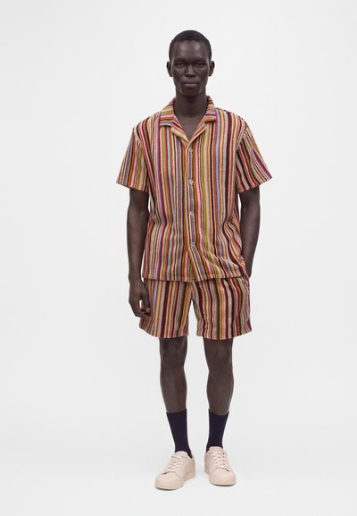 Homme portant une chemise à manches courtes rayée verticale multicolore et un short assorti, des baskets claires et des chaussettes foncées, debout devant un fond blanc.