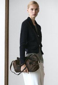 Donna con capelli biondi che indossa una giacca nera lavorata a maglia e pantaloni bianchi, che tiene una borsa in pelle marrone scuro per la tracolla al suo fianco.