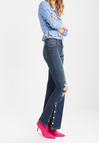Jeans a vita alta, in denim scuro, a campana con toppe strappate, presentano spacco laterale e dettagli con bottoni, abbinati a una camicetta floreale blu e tacchi rosa.