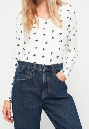 Femme portant une chemise blanche à manches longues avec de petits motifs floraux noirs et un jean bleu taille haute, une main dans la poche.