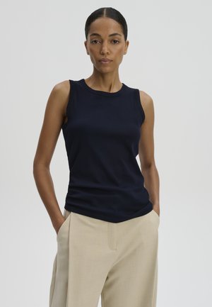 Femme aux cheveux foncés attachés, portant un haut noir sans manches et un pantalon beige, debout les mains dans les poches devant un fond uni.
