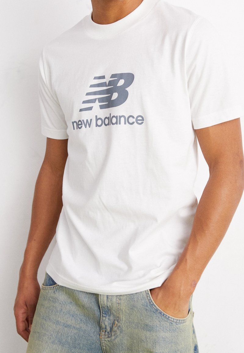 Bílý bavlněný tričko s velkým šedým logem "NB" a textem "new balance". Kulatý výstřih, krátké rukávy, volný střih.