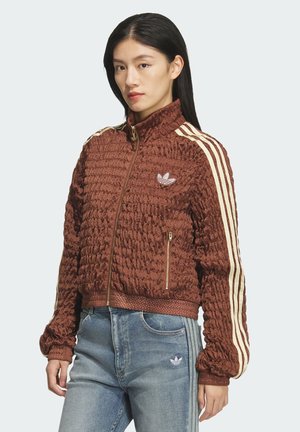 Ženska v rjavi teksturirani jakni z zadrgo, s kremnimi črtami na rokavih in svetlo modrimi kavbojkami, stoji pred enobarvnim ozadjem.