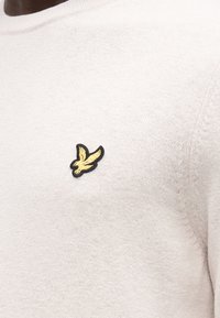 Närbild av beige stickad tröja med liten gul fågelmärke på bröstet, som visar stickningens struktur och sömdetaljer.