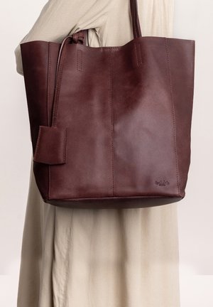 Brown leather tote bag with sturdy handles, smooth texture, and a side pocket. Minimalist design with visible stitching and an embossed logo detail.