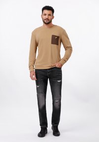 Beige long-sleeve shirt with brown patch pocket and embroidered accent, paired with black ripped jeans and black sneakers.