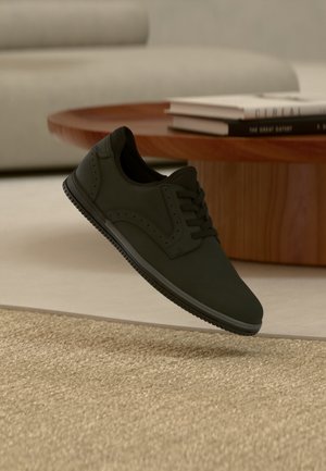Czarny casualowy sneaker unoszący się nad teksturowanym beżowym dywanem z okrągłym drewnianym stolikiem kawowym i książkami w tle.
