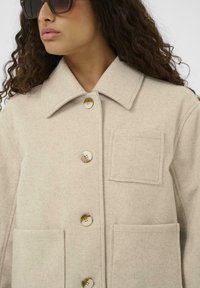 Manteau en laine mélangée beige avec un col pointu, présentant de grands boutons et une poche poitrine. Texture douce et design ajusté.