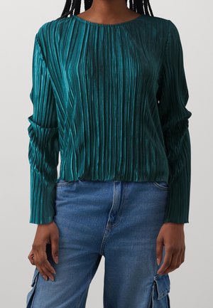 Kvinde iført en langærmet, mørk teal, lodret plisseret top kombineret med mellemblå denim cargo bukser.