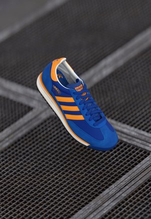 Niebiesko-pomarańczowy but sportowy Adidas z trzema paskami i sznurowadłami, unoszący się nad metalową kratownicą na podłodze w miejskim otoczeniu.