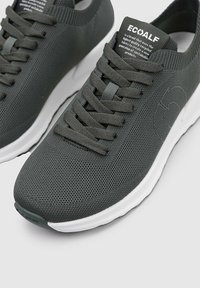 Zapatilla de deporte gris oscuro con parte superior de malla texturizada, cordones planos y una suela blanca con un logotipo prominente en el costado.