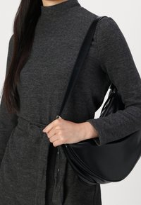Schwarze Leder-Schultertasche mit geschwungenem Design, glatter Textur und verstellbarem Tragegurt, gehalten von einer Person in einem grauen Langarmkleid.