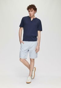 Marineblaues Kurzarmhemd mit V-Ausschnitt und Knöpfen, kombiniert mit hellblauen Shorts mit umgeschlagenen Säumen und weißen Sneakers mit hellbraunen Sohlen.