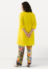 Tunic di lino gialla con maniche lunghe, abbinata a pantaloni colorati con un motivo di frutta e edifici, indossata con tacchi bianchi con cinturini.