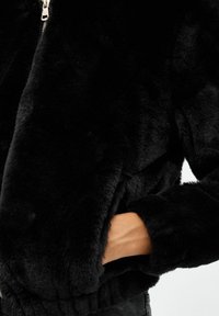 Chaqueta de piel sintética negra con cierre de cremallera en la parte delantera, dobladillo elástico y dos bolsillos laterales. Suave al tacto y con un acabado lujoso que realza su apariencia.