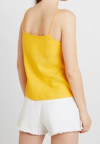 Donna che indossa un top giallo senza maniche con spalline sottili e pantaloni denim bianchi con orli sfrangiati, vista da dietro su uno sfondo semplice.