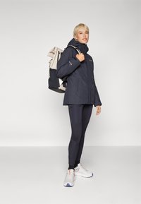 Dark blue waterproof jacket with a hood, paired with black leggings and white athletic shoes. Carrying a light-colored backpack.