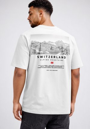 Man wearing white t-shirt with black and white Swiss alpine mountains photo and text on the back, including coordinates and facts.