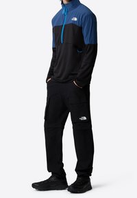 Black and blue zip-neck pullover with a textured pattern, paired with black cargo pants featuring reflective logo accents and multiple pockets.