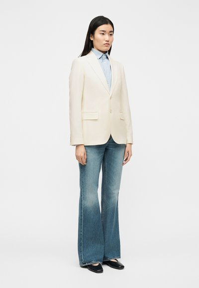 Γυναίκα όρθια με εκρού σακάκι πάνω από γαλάζιο πουκάμισο με κουμπιά, μπλε τζιν καμπάνα και μαύρα μυτερά παπούτσια σε λευκό φόντο.