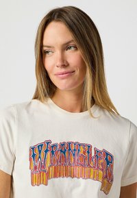 Licht beige katoenen t-shirt met een kleurraf, retro-stijl "WRANGLER" graphic in blauw, rood en geel. Korte mouwen, ronde halslijn.