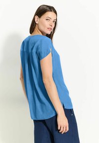 Blaues T-Shirt mit kurzen Ärmeln, weitem Schnitt, weichem Material und einem Faltendetail am Rücken. Seitenschlitze erhöhen die Luftzirkulation und den Komfort.