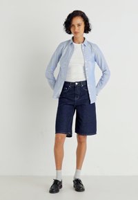 Lyseblå stripete skjorte med knapper over en hvit t-skjorte, kombinert med mørk denimshorts og svarte sko. Korte ermer og ledig passform.