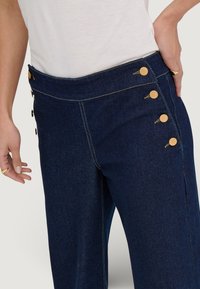 Donker denimbroek met een hoge tailleband, voorzien van vijf decoratieve gouden knopen aan de linkerkant en contrasterende stikseldetails.
