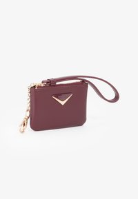 Piccola pochette color maroon con zip dorata, emblema triangolare dorato e catena dorata attaccata con chiusura su sfondo bianco.