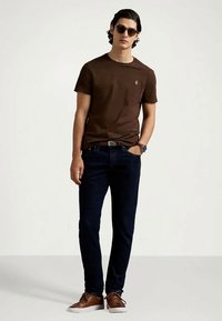 Καφέ βαμβακερό μπλουζάκι με μικρό λογότυπο στο στήθος, συνδυασμένο με σκούρες τζιν και καφέ παπούτσια. Το μοντέλο φορά γυαλιά ηλίου και στέκεται με χαλαρό τρόπο.