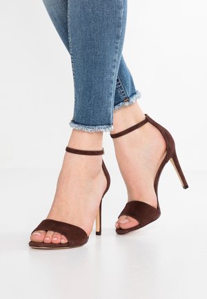 Brązowe zamszowe sandały na wysokim obcasie z paskami w okolicy kostki i otwartym czubkiem. Posiadają smukły obcas i gładką cholewkę. Noszone z dżinsami w niebieskim kolorze z postrzępionymi nogawkami.