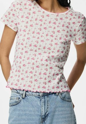 Mujer con una camiseta blanca de manga corta con un pequeño estampado floral rosa y bordes ondulados, combinada con vaqueros denim azul claro.