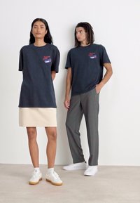 Dos modelos están de pie uno al lado del otro. La modelo femenina lleva una camiseta oversized de color gris oscuro y una falda beige; el modelo masculino lleva una camiseta a juego y pantalones grises.