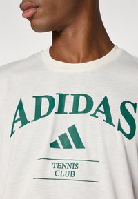 Camiseta branca de algodão com texto e logótipo "ADIDAS" em verde; decote redondo; apresenta "TENnis CLUB" em letras menores por baixo.