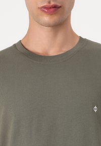 Olivgrünes Baumwoll-T-Shirt mit Rundhalsausschnitt, strukturiertem Finish und einem kleinen weißen Logo-Akzent auf der linken Seite.