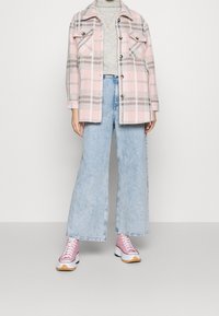 Veste oversize à carreaux roses, pull gris, jean large bleu clair, et baskets roses avec des détails blancs et beige. Look décontracté en couches.