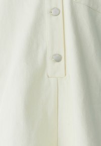 Lichtgele kledingstuk van een gestructureerde stof, voorzien van zilveren knopen en een subtiele plooienontwerp. Close-up weergave waarbij de knoopdetails worden benadrukt.