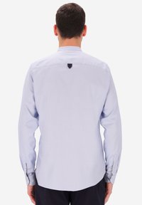 Uomo di spalle che indossa una camicia a maniche lunghe azzurro chiaro con polsini abbottonati e pantaloni scuri, su uno sfondo bianco semplice.