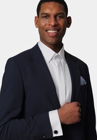 Navy blue suit jacket with a textured finish, white dress shirt, and contrasting cufflink. White pocket square with a subtle check pattern.