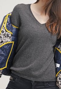 Szary sweter z dekoltem w serek w miękkim materiale, noszony pod niebieską pikowaną kurtką w kwiatowy wzór z żółtymi wykończeniami.