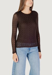 Top lungo in pizzo nero con maniche lunghe, caratterizzato da un motivo a zigzag, scollatura rotonda e una texture leggermente trasparente, abbinato a jeans azzurri chiari.