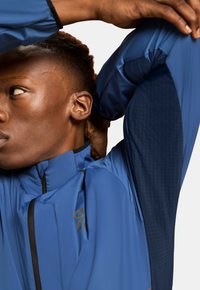 Blue jacket with a zip, featuring a mixed texture design and navy sleeves. Details include a logo on the chest and a high collar for coverage.