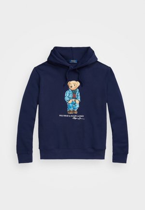 Sudadera con capucha azul marino con un gráfico de un oso que lleva una chaqueta vaquera y jeans, con el texto "Polo Bear by Ralph Lauren" debajo.
