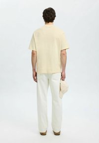 Jasno beżowa koszula z krótkim rękawem, bawełniany materiał, luźny krój. Połączona z białymi spodniami o szerokich nogawkach i trzymany beżowy kapelusz w pasującym kolorze.