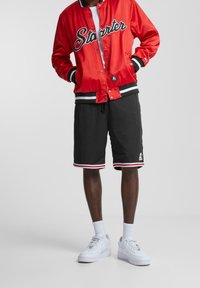 Rød satin varsityjakke med sorte og hvide accenter, parret med sorte shorts med rød kant og hvide sneakers.