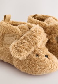 Puhasti bež copati v obliki psa, z teksturiranim materialom, povešenimi ušesi in majhnimi črnimi očmi. Vključuje tkaninski zanko za enostavno prijemanje.