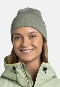 Gorro de punto acanalado verde claro con un pequeño parche de logo. Usado con una chaqueta verde clara que cuenta con un collar y cremallera amarillos.