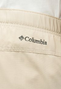 Εγγύς άποψη μπεζ υφασμάτινης ζώνης μέσης με ελαστική ταινία και μαύρο κεντημένο λογότυπο Columbia.