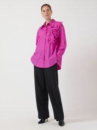 Camisa rosa brillante de gran tamaño con un detalle de aplique floral texturizado en el hombro izquierdo, combinada con pantalones de pierna ancha negros y tacones.